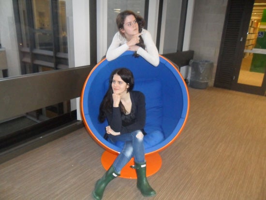 One girl sitting in a wombchair, contemplating with her fist raised to her chin. Another girl stands behind the wombchair with her arms resting over the top, pointing at something out of sight