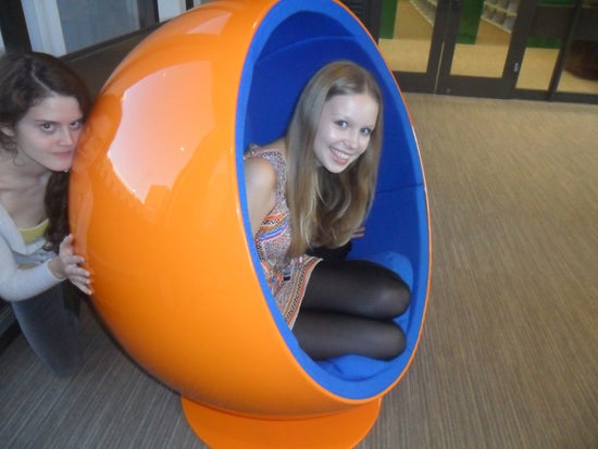 One girl sitting in the wombchair and peeking out to smile at the camera. Another girl can be seen kneeling behind the wombchair with her hands on the back of it
