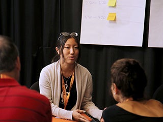 A student explains something to 2 staff members at a table. In the background is a board with sticky notes on it.