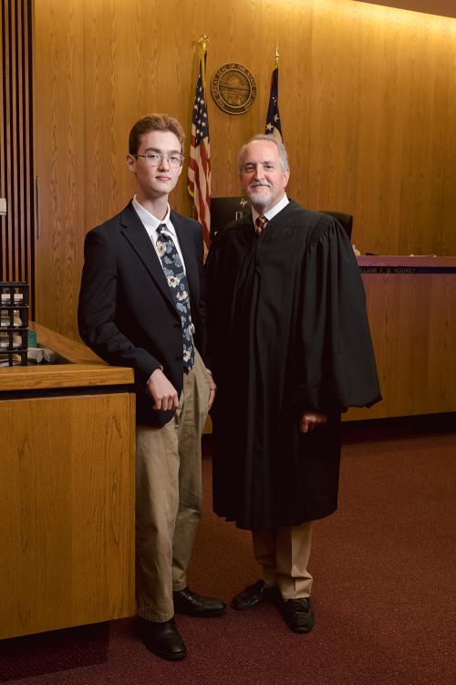 a student wearing a suit coat and tie stands next to a judge wearing robes in a courtroom