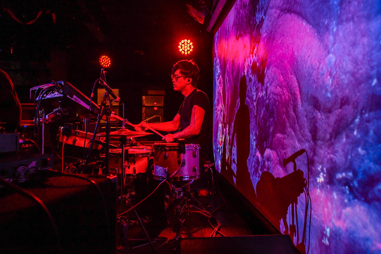 A drummer performs against a multicolored backlit screen.