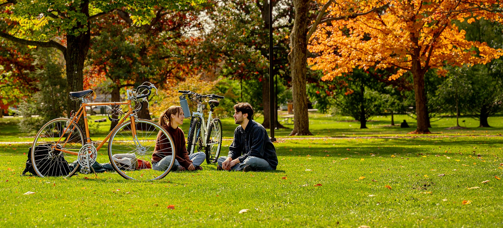 Two students sit on a green lawn with an autumnal backdrop; two bicycles stand nearby.