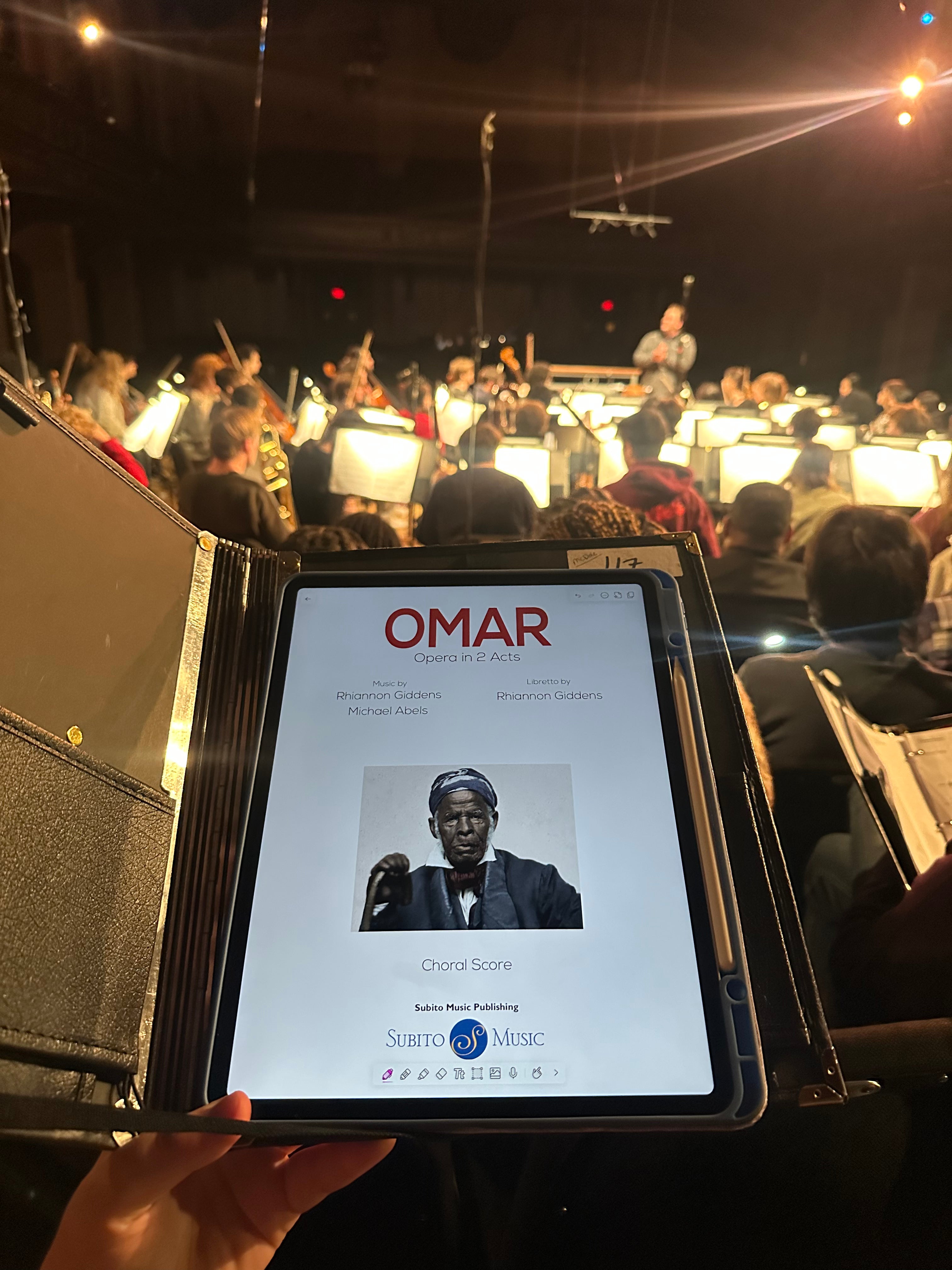 Rebekah holding up her score onstage at Finney Chapel in front of the Oberlin Orchestra in a dress rehearsal for Omar.