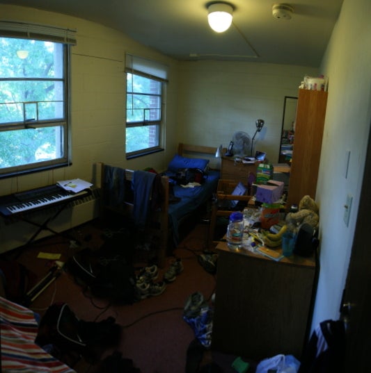 A messy dorm room with clothes on the floor and cluttered surfaces