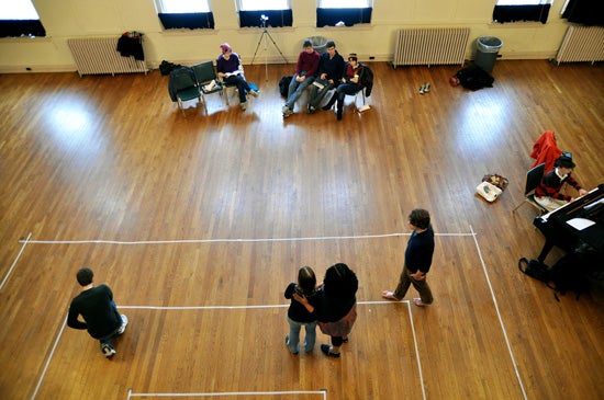 Performers rehearse in a large room with a wood floor, which has sections marked ou with tape.