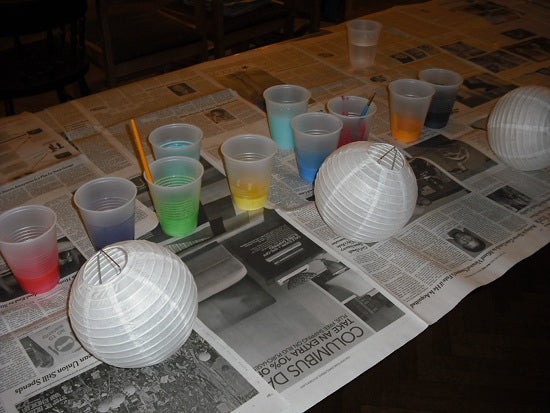 Unpainted lanterns on newspaper next to cups of colorful paint.