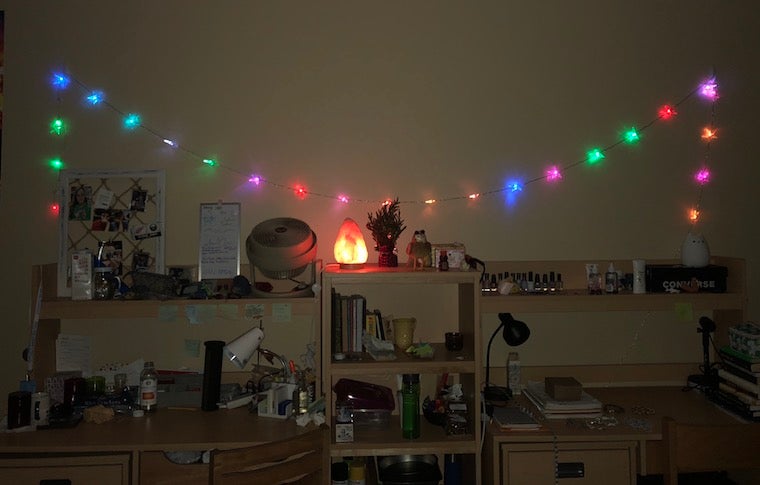 Two desks, side by side, decorated with many trinkets, and a bookshelf, also very decorated, between them. A string of colorful lights hangs above the desks, and a salt lamp shines on top of the bookshelf.