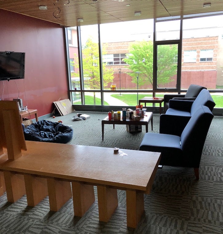 A dorm lounge, featuring chairs, tables, a TV, and a large window. Through the window, trees and the side of another building can be seen.