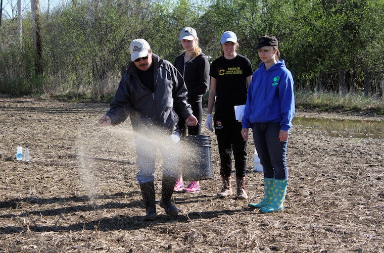 Three students look on as a man throws seeds onto a muddy field