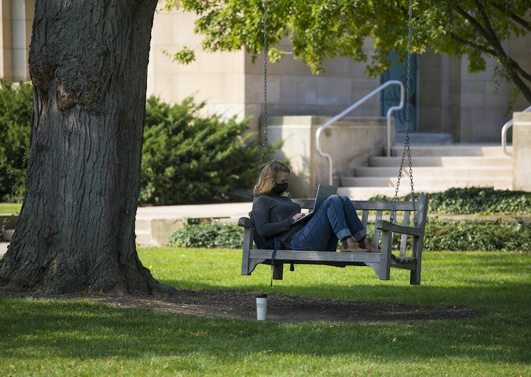 A student types on a laptop while sitting in a tree swing.