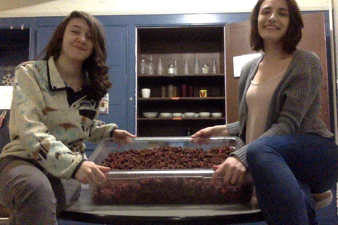 Megan and Lily sit on the tasty things table in Fairchild, holding their uncovered container of granola and tilting it toward the camera. They are smiling.