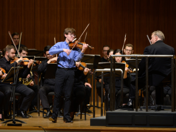 young violinist in blue shirt playing onstage with orchestra and conductor