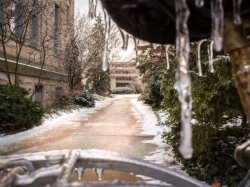 An icicle hangs from a bike seat in the foreground. Behind, a snowy path leads to a building.
