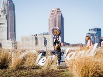 Photograph of a city backdrop, a sign that says, "Cleveland," and three women posing by the sign.