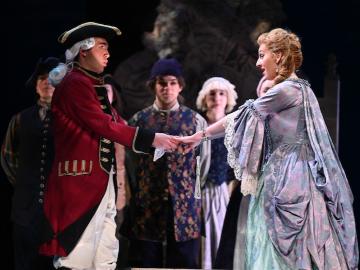 Two actors dressed in 18th century costume hold hands on stage.
