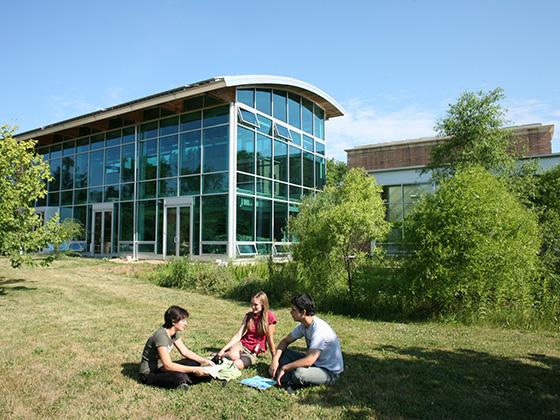 On a sunny day, students sit in the grass in front of the glass facade of the Lewis Center.