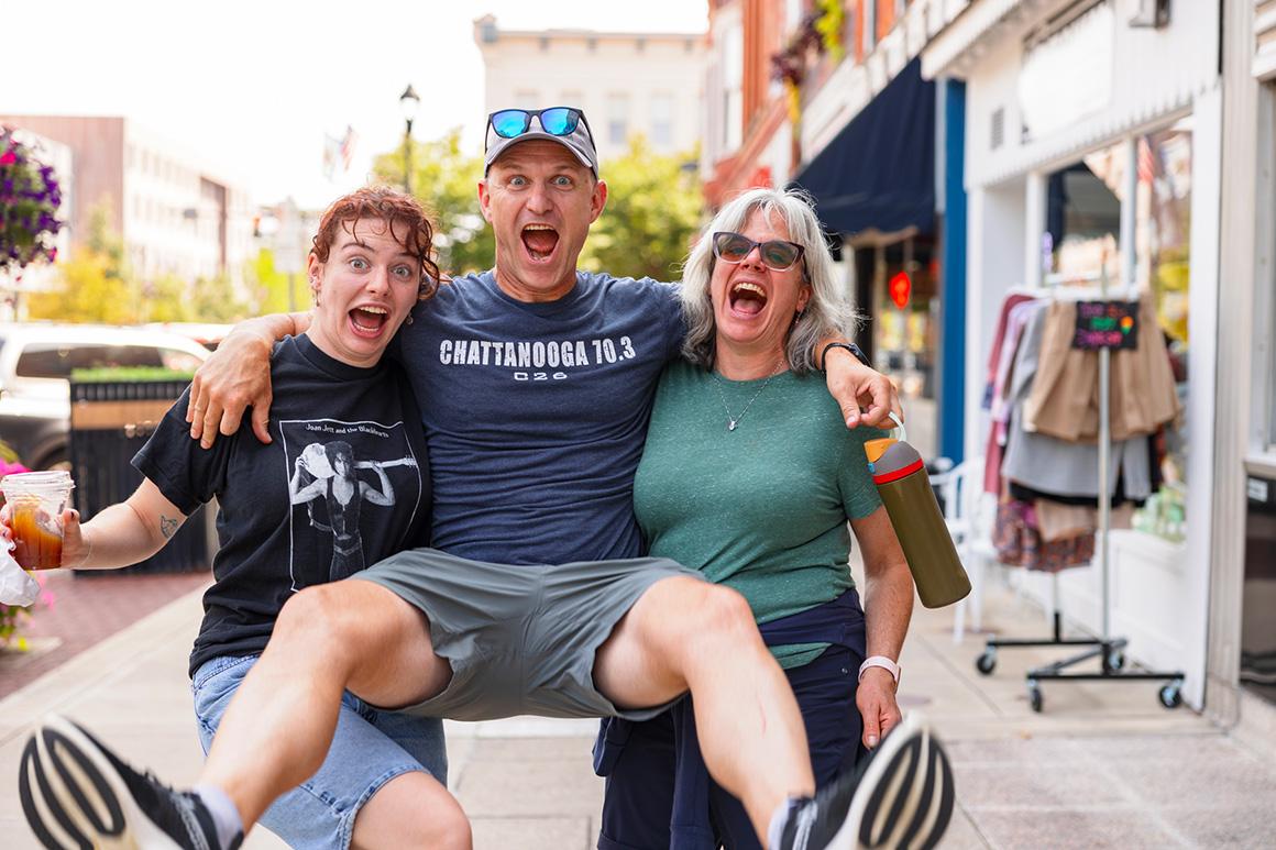 On a downtown Oberlin street, a student and her mom hold up her dad, whose legs are stretched in front of him off the ground. They are all making silly faces and having a great time.