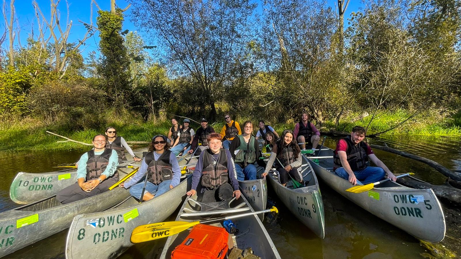 People in about a dozen canoes clustered together, all facing forward, with a riverbank behind them.