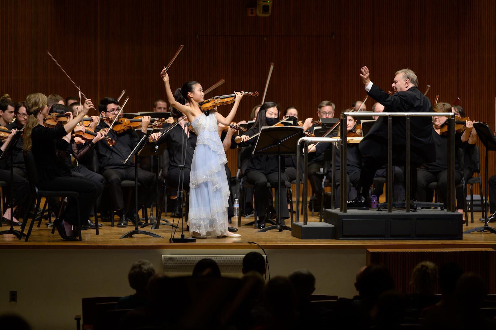 violin soloist in blue dress performs on stage with orchestra and conductor