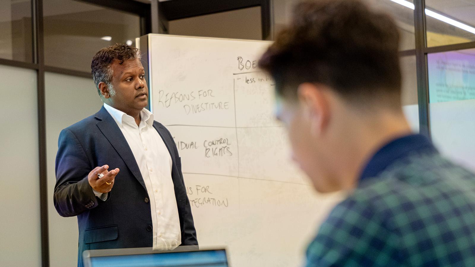 A professor speaks while a student takes notes on a laptop.