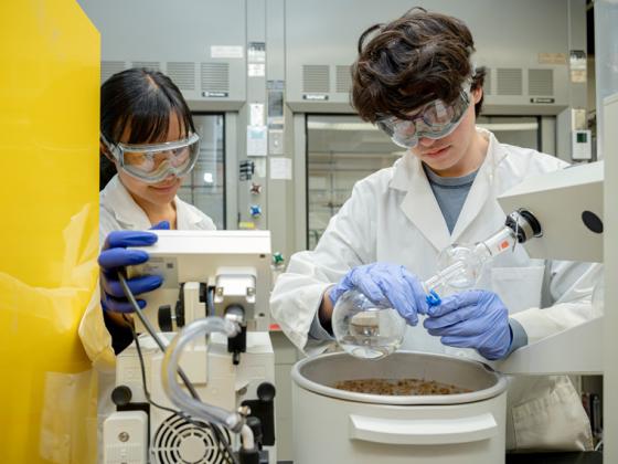 Two people wearing protective gear like googles and gloves conduct an experiment in a lab