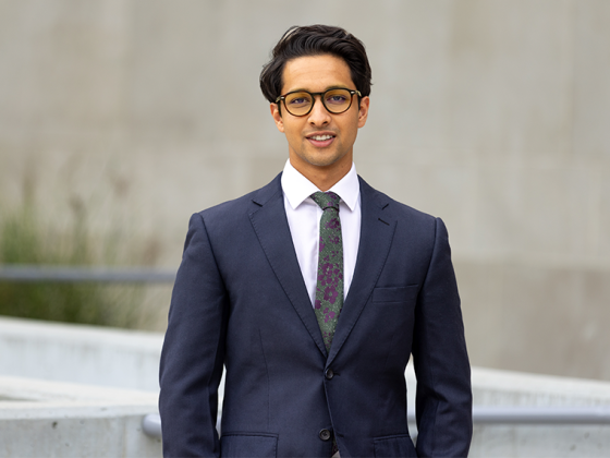 Student with glasses in suit smiles at camera in front of tan building.