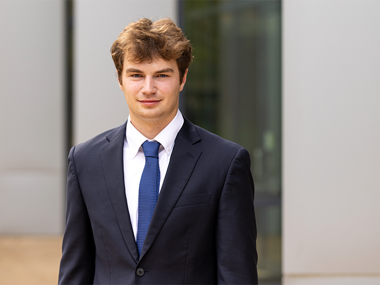 Student in blue suit poses in front of steel and glass building.