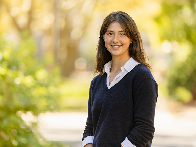 Student smiles against fall foliage campus background.