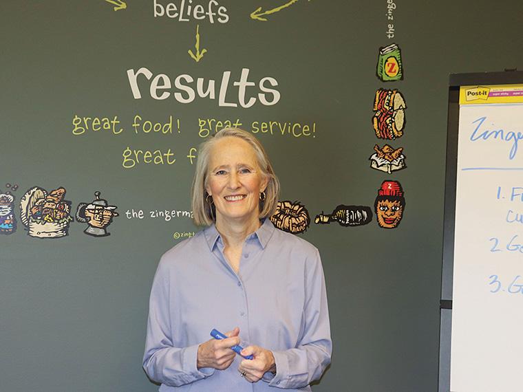 Hanna Raskin standing in front of a chalkboard, which has a border of colorful drawings of food and people.