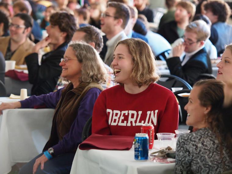 People at a large gathering, seated at tables with tablecloths. Most are smiling.